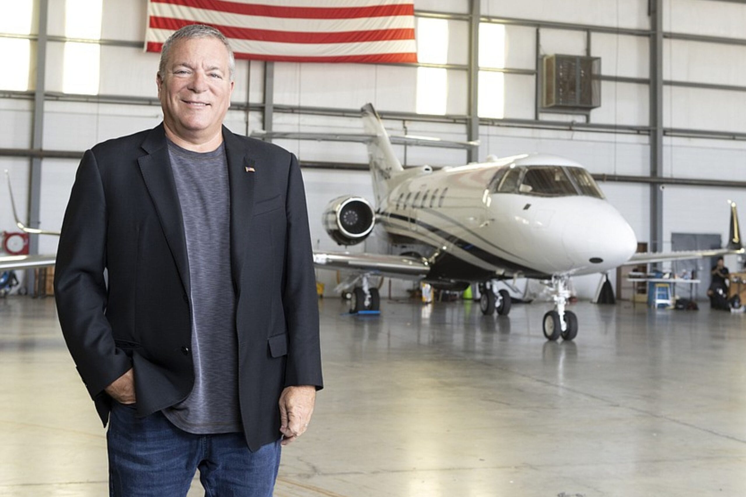 A man in a black blazer and gray shirt stands smiling in an aircraft hangar with a private jet behind him and an American flag hanging on the wall. South Florida Business & Wealth
