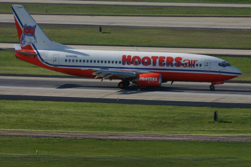 A Hooters Air passenger airplane painted in white and orange with owl logo on the tail sits on a runway surrounded by grassy areas at an airport. South Florida Business & Wealth
