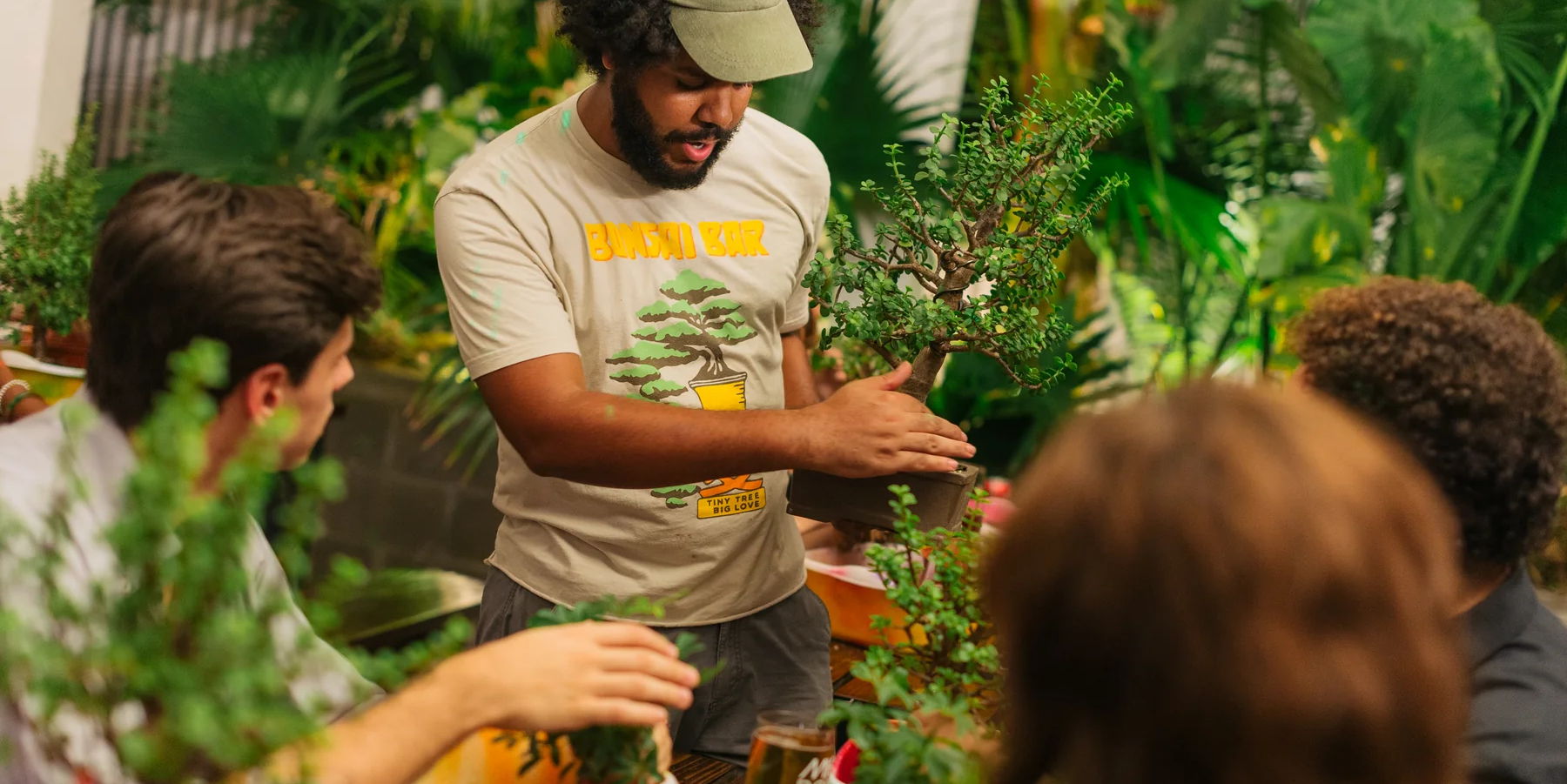 A man wearing a cap and a "Bonsai Bar" t-shirt demonstrates bonsai techniques to three people seated around a table covered with small bonsai trees, surrounded by lush green plants. South Florida Business & Wealth