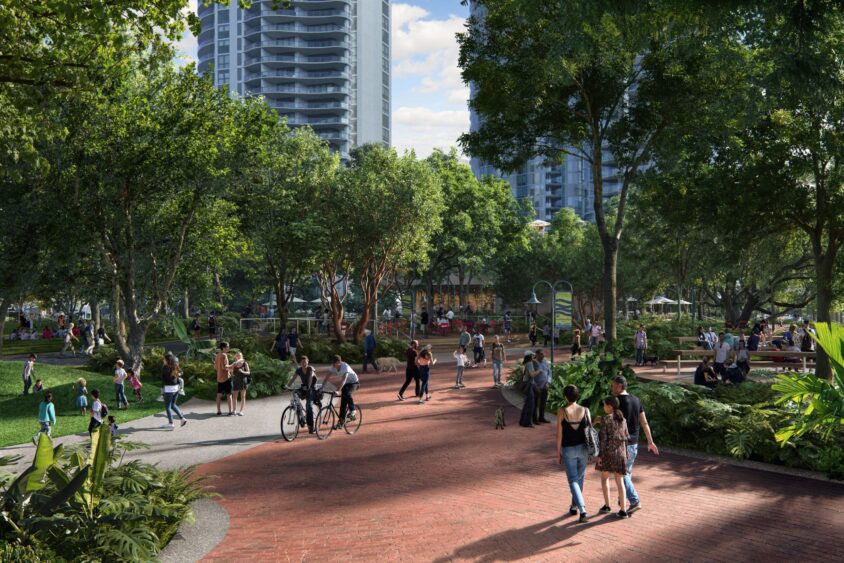A lively city park with many people walking, biking, and relaxing among trees and gardens, with tall modern buildings in the background on a sunny day. South Florida Business & Wealth