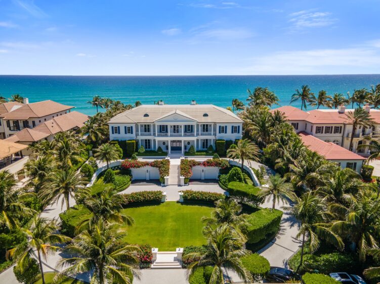 Aerial view of a large, elegant white mansion with manicured gardens and palm trees, located on a beachfront with clear blue ocean and sky in the background. Neighboring luxurious homes line both sides. South Florida Business & Wealth
