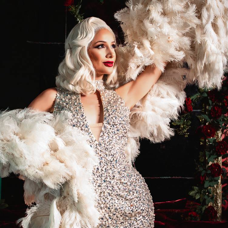 A glamorous woman in a sparkling, silver sequin dress poses with white feathered fans. She has platinum blonde, wavy hair, bold red lipstick, and is set against a dark background decorated with red roses. South Florida Business & Wealth