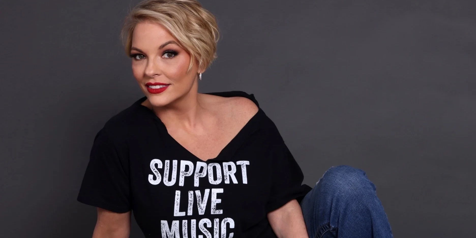 A woman with short blonde hair and red lipstick sits against a gray background, wearing a black off-shoulder T-shirt that says "SUPPORT LIVE MUSIC" and blue jeans. She is smiling and looking at the camera. South Florida Business & Wealth