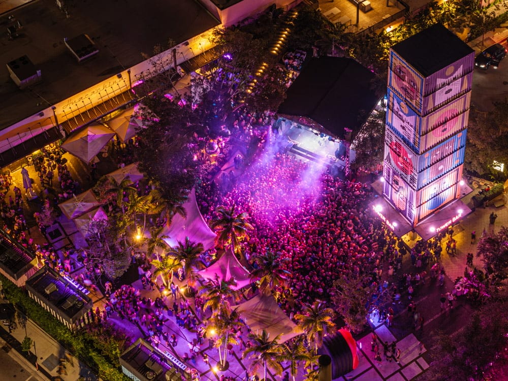 Aerial view of a vibrant outdoor music festival at night, with a large crowd gathered around a stage, colorful lighting, palm trees, tents, and a tall tower made of stacked shipping containers. South Florida Business & Wealth