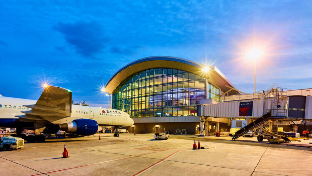 A Delta airplane is parked at a brightly lit airport terminal at dusk, with the modern glass building illuminated and ground service vehicles nearby. South Florida Business & Wealth