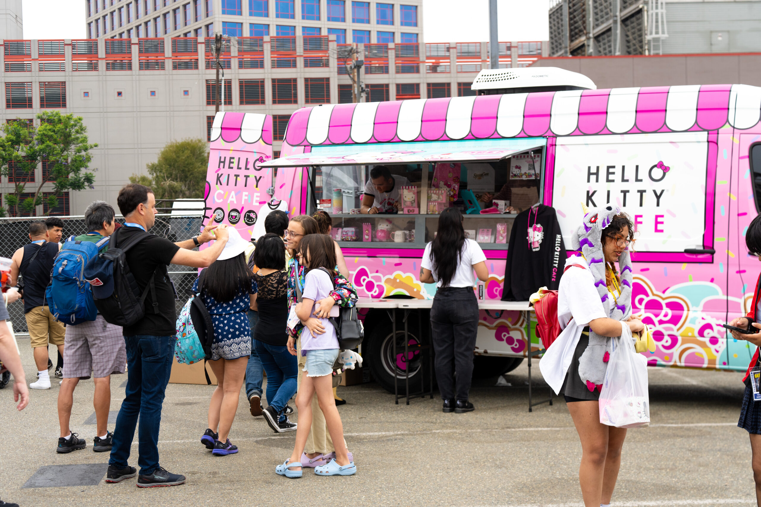 A group of people line up at a pink Hello Kitty Cafe food truck, decorated with floral and cartoon designs, in an outdoor urban area on a cloudy day. Some people are ordering, while others stand nearby or carry treats. South Florida Business & Wealth
