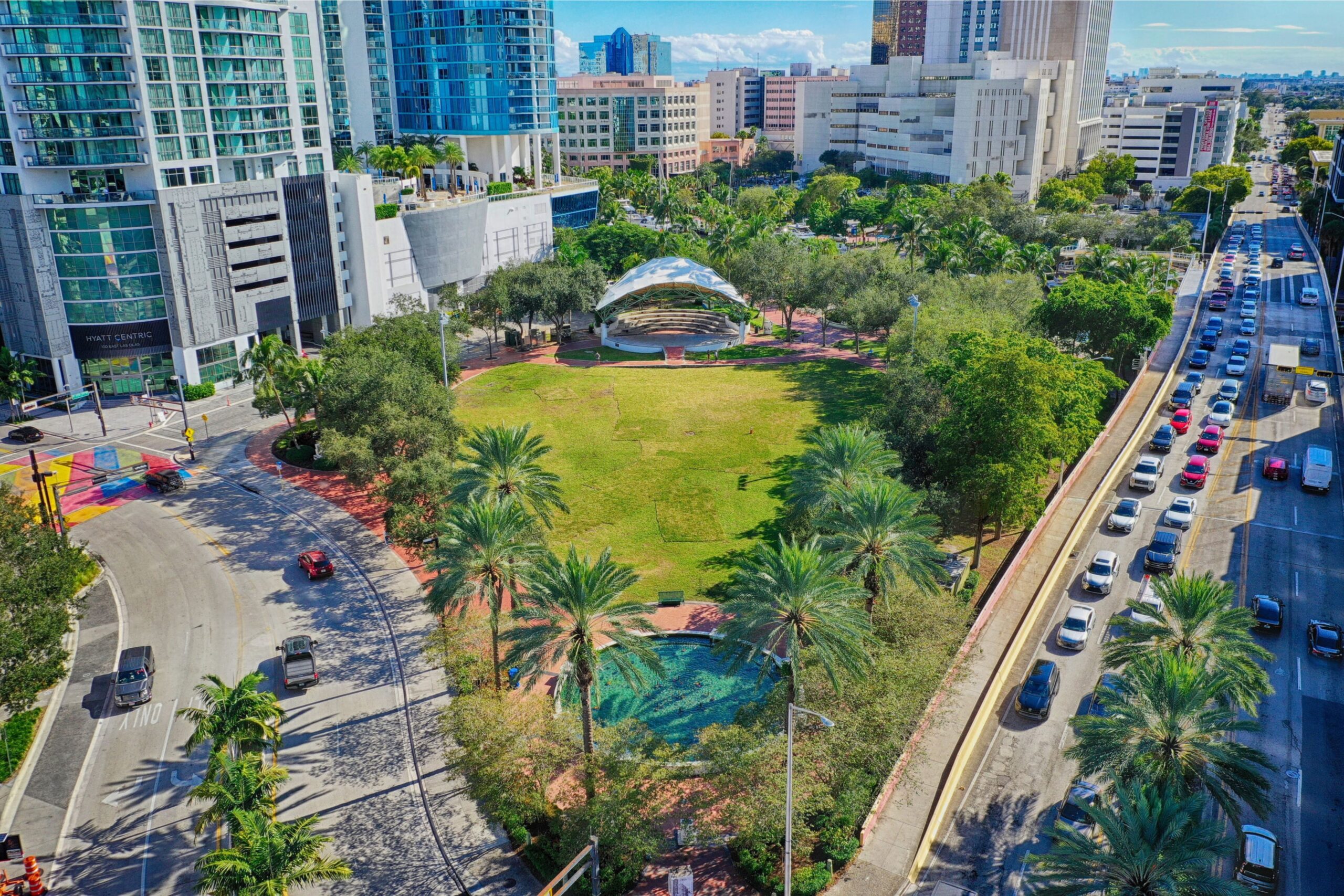 Aerial view of a green city park surrounded by tall buildings and busy roads with cars, palm trees, an outdoor stage, and a small pond under a clear blue sky. South Florida Business & Wealth