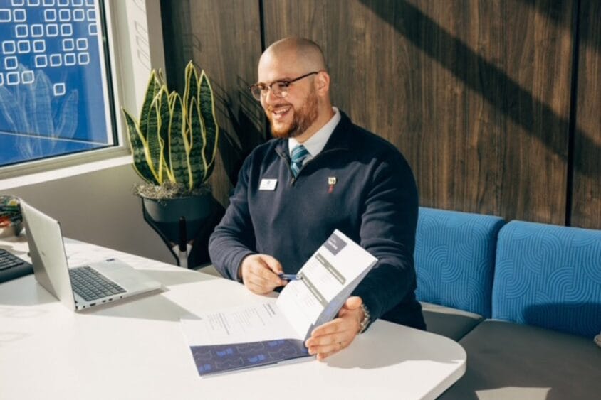 A smiling man wearing glasses and business attire sits at a desk with a laptop, holding and presenting a document in a bright office with a plant and blue seating in the background. South Florida Business & Wealth