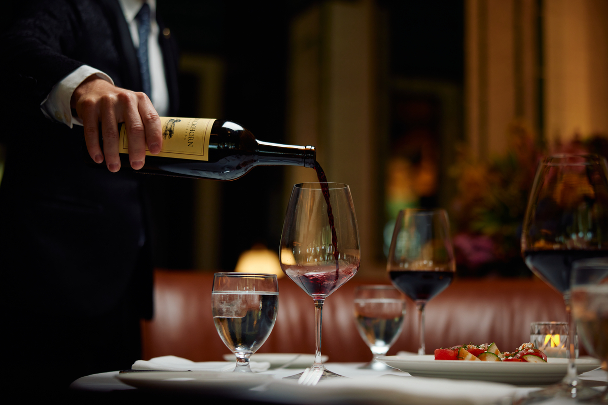 A person in a suit pours red wine from a bottle into a wine glass at a restaurant table, which is set with water glasses, more wine glasses, and plates of food. The setting appears elegant and dimly lit. South Florida Business & Wealth