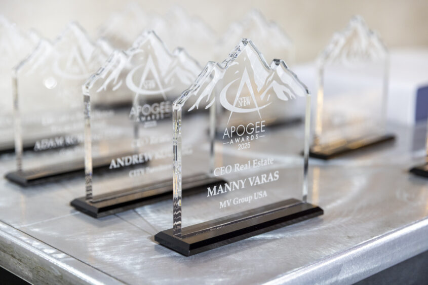 A close-up of several clear glass awards shaped like mountain peaks, engraved with "Apogee Awards 2025" and recipient names, displayed on a silver table. South Florida Business & Wealth