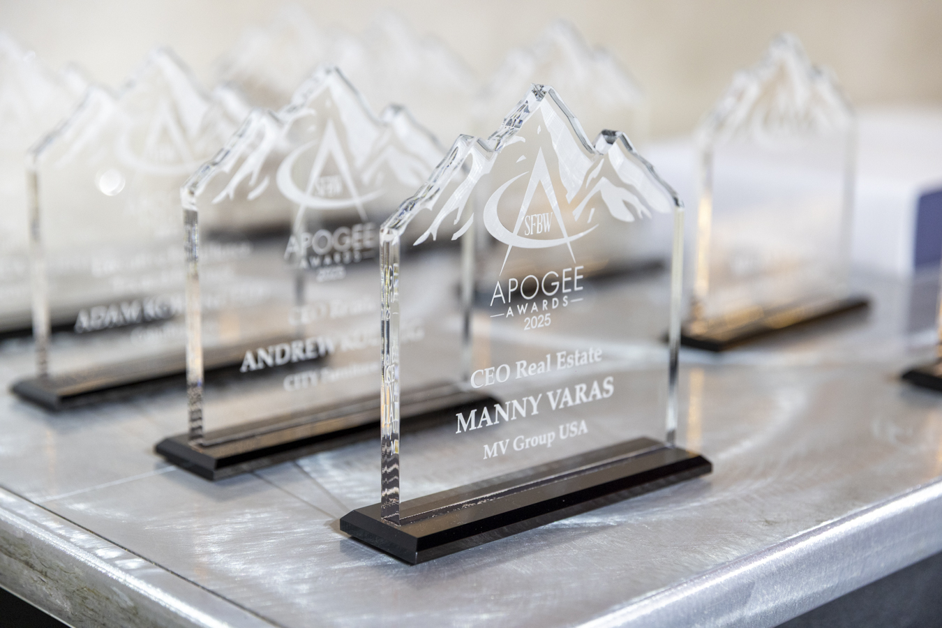 A close-up of several clear glass awards shaped like mountain peaks, engraved with "Apogee Awards 2025" and recipient names, displayed on a silver table. South Florida Business & Wealth