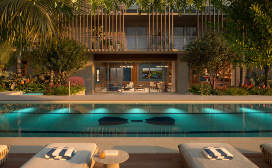 A serene outdoor pool with lounge chairs and towels, surrounded by lush greenery, in front of a modern building with balconies and large glass doors leading to a well-lit interior. South Florida Business & Wealth