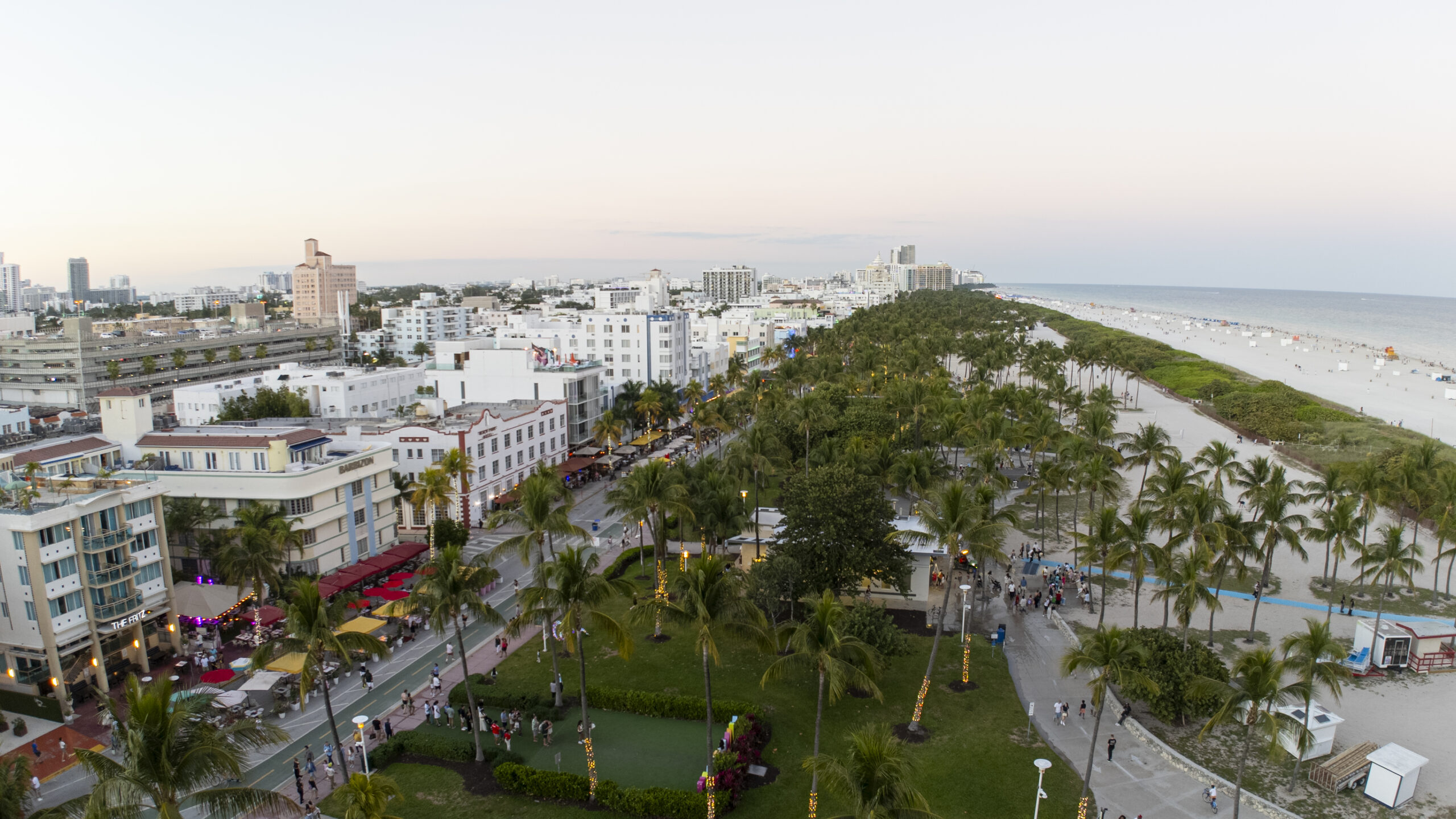 Aerial view of a city beachscape with palm trees, green park, busy streets, white Art Deco buildings, and a sandy beach stretching along the ocean under a clear sky. South Florida Business & Wealth
