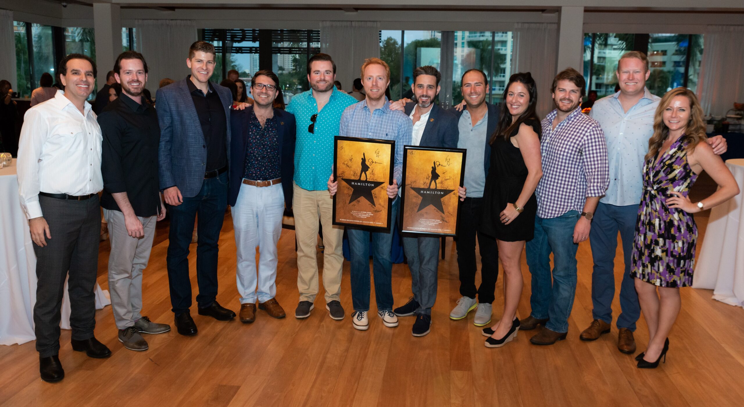 A group of twelve people, including men and women, stand indoors on a wooden floor, smiling at the camera. Two men in the center hold framed "Hamilton" posters. The group appears to be at a celebratory event. South Florida Business & Wealth