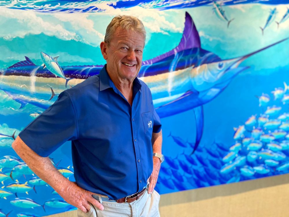 An older man in a blue shirt stands smiling in front of a colorful mural featuring a large marlin and a school of fish in a vibrant underwater scene. South Florida Business & Wealth