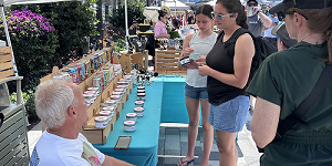 People browse and purchase items from a vendor at an outdoor market stall displaying colorful canned goods and jars on a turquoise tablecloth. South Florida Business & Wealth