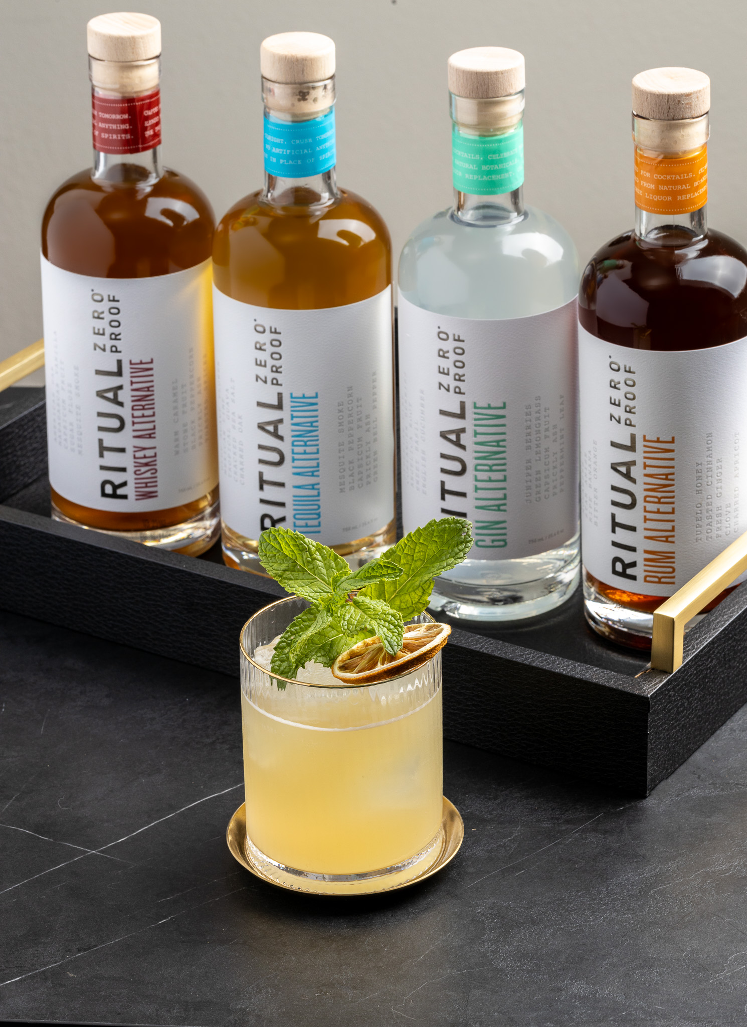 A cocktail garnished with mint and dried citrus sits on a gold coaster in front of four Ritual Zero Proof bottles, displayed upright in a black tray on a dark surface. South Florida Business & Wealth