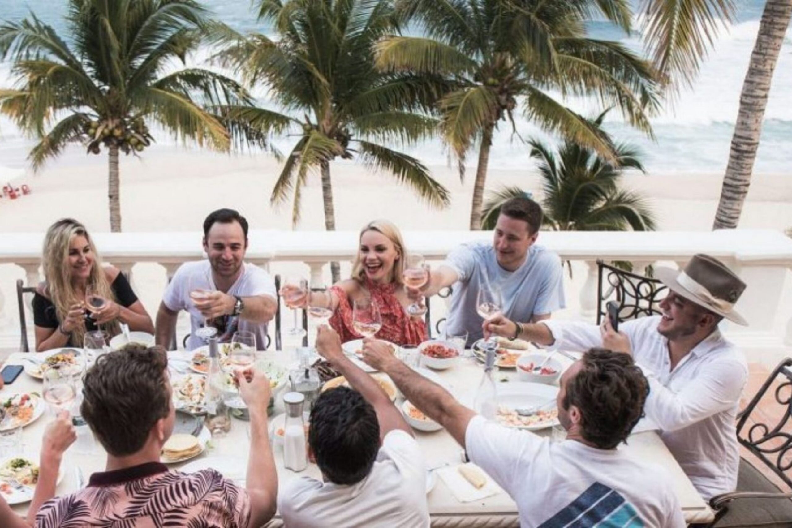 A group of eight people sit around an outdoor table, raising glasses in a toast. The table is set with food, and palm trees and an ocean view are in the background, creating a festive and tropical atmosphere. South Florida Business & Wealth