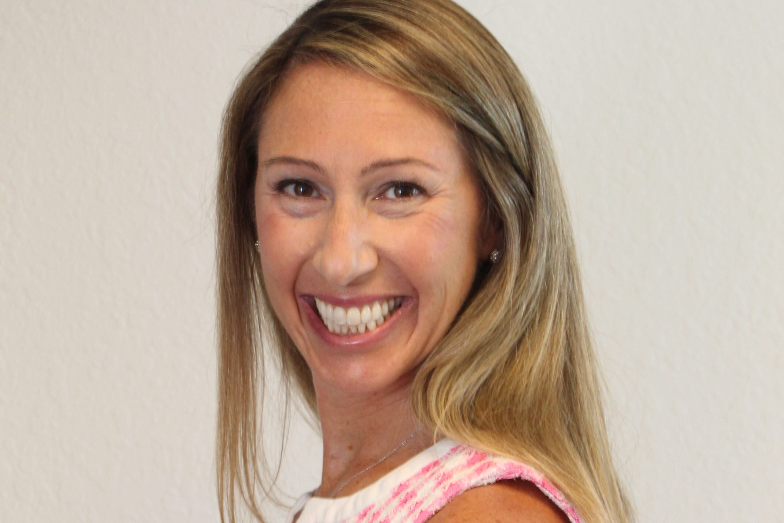 A woman with long, straight blonde hair smiles broadly at the camera. She is wearing a sleeveless pink and white top and is posed in front of a plain, light-colored background. South Florida Business & Wealth
