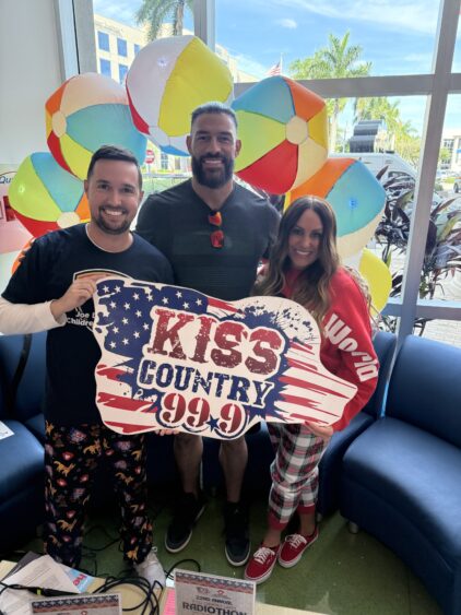 Three people smiling and holding a KISS Country 99.9 sign indoors, with colorful beach balls and palm trees visible through the window behind them. Two are wearing pajama pants; festive decorations create a cheerful atmosphere. South Florida Business & Wealth