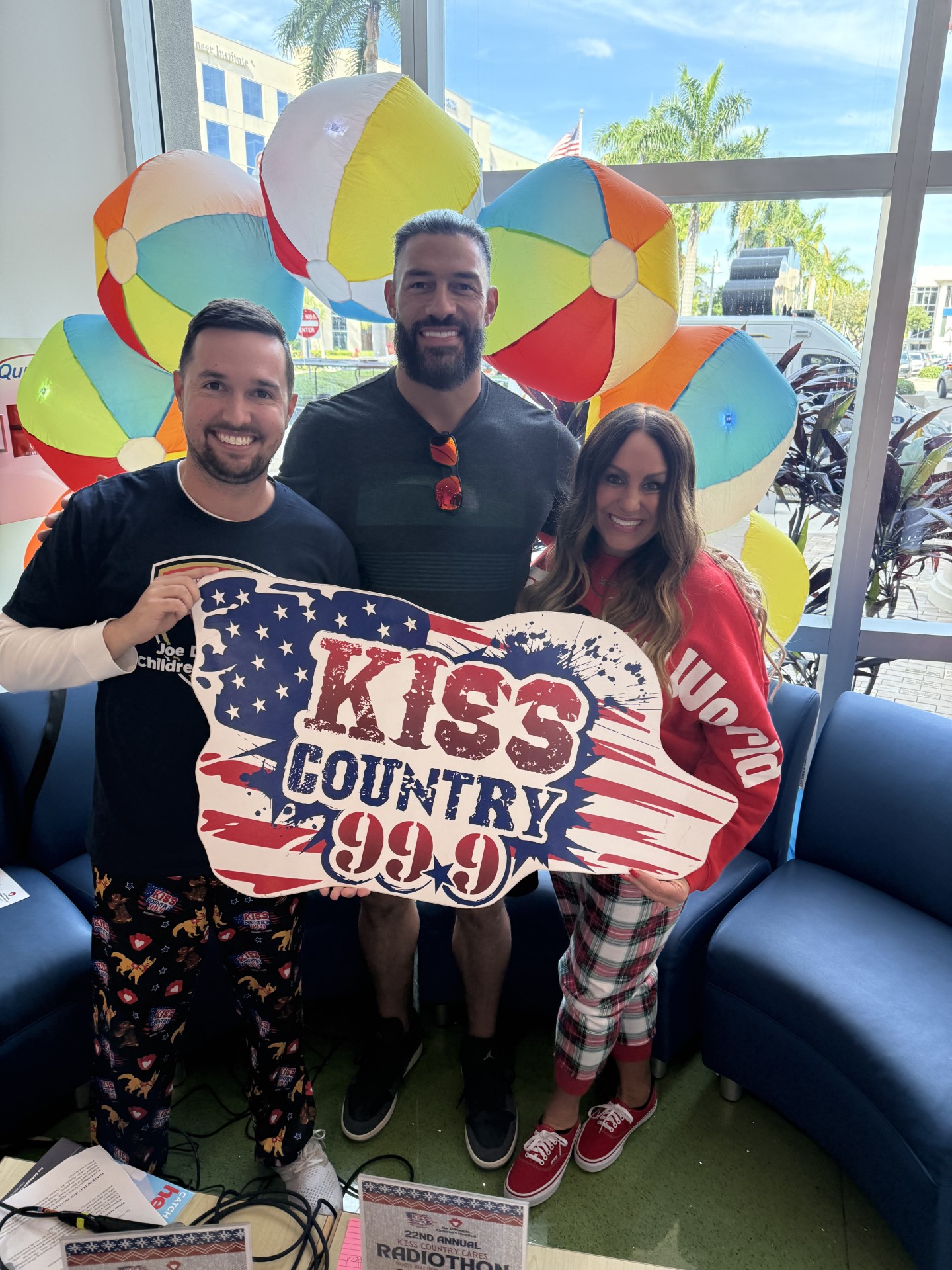 Three people smiling and holding a KISS Country 99.9 sign indoors, with colorful beach balls and palm trees visible through the window behind them. Two are wearing pajama pants; festive decorations create a cheerful atmosphere. South Florida Business & Wealth