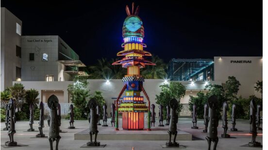A tall, colorful, illuminated sculpture stands in an outdoor plaza at night, surrounded by dark human-like statues. Modern buildings and lush greenery are visible in the background, with brand signage on the walls. South Florida Business & Wealth