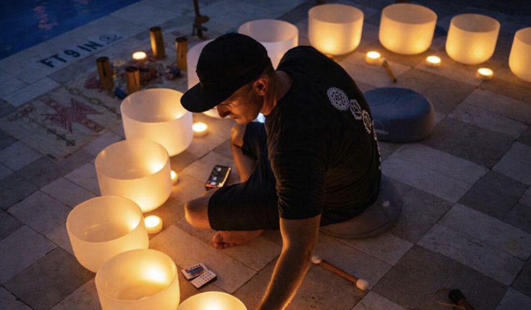 A person sits cross-legged on a tiled floor, surrounded by glowing candles and white crystal singing bowls near a pool at night, preparing for a sound healing session. South Florida Business & Wealth