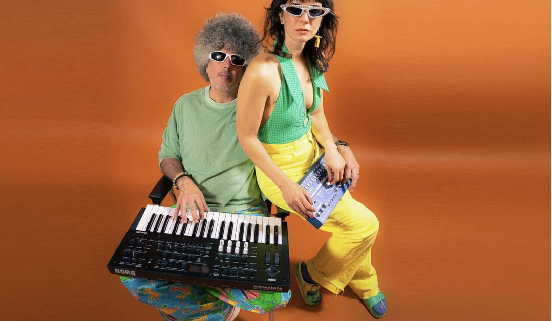 Two people pose against an orange background. One sits with a Korg keyboard, wearing a green shirt, blue pants, and white sunglasses. The other stands, wearing a green top, yellow pants, white sunglasses, and holding a small sound mixer. South Florida Business & Wealth