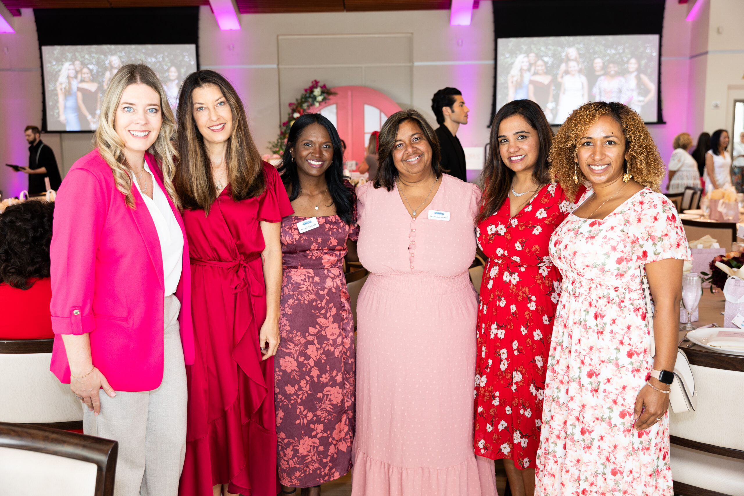 Six women stand together, smiling at the camera. They are dressed in various shades of pink, red, and white. The background shows a decorated event space with tables, chairs, and large screens displaying group photos. South Florida Business & Wealth