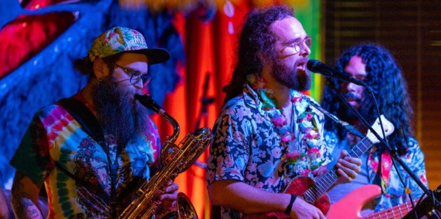 Three musicians wearing colorful shirts perform on stage; one plays saxophone, one sings and plays guitar, and another plays bass. The background features vibrant, colorful decorations and lighting. South Florida Business & Wealth