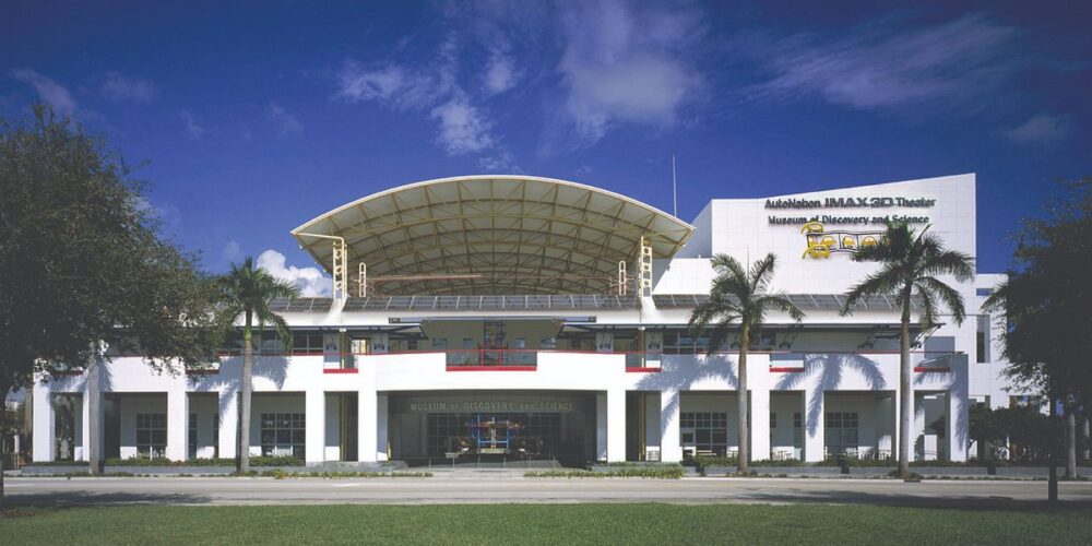 A white modern building with palm trees in front, labeled "Museum of Discovery and Science," featuring a distinctive arched roof and banners, under a blue sky with scattered clouds. South Florida Business & Wealth