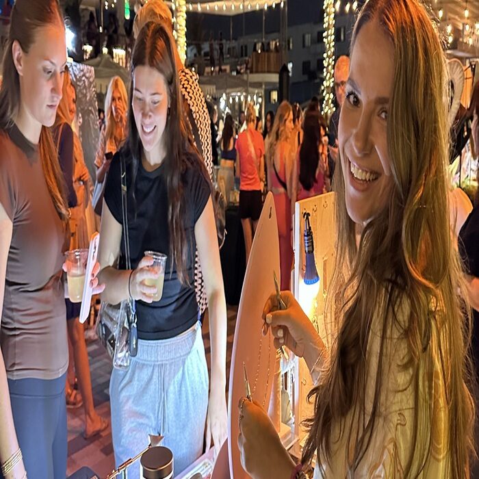 Three young women enjoy an outdoor night market, smiling and holding drinks. One woman in the foreground grins at the camera while working on a craft at a booth. Warm lights and a lively crowd fill the festive atmosphere. South Florida Business & Wealth