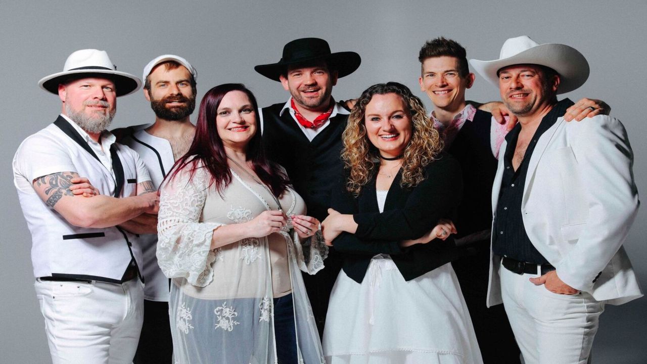 A group of seven people stand together smiling, wearing a mix of white and black outfits, including hats, jackets, and dresses, against a plain grey background. South Florida Business & Wealth