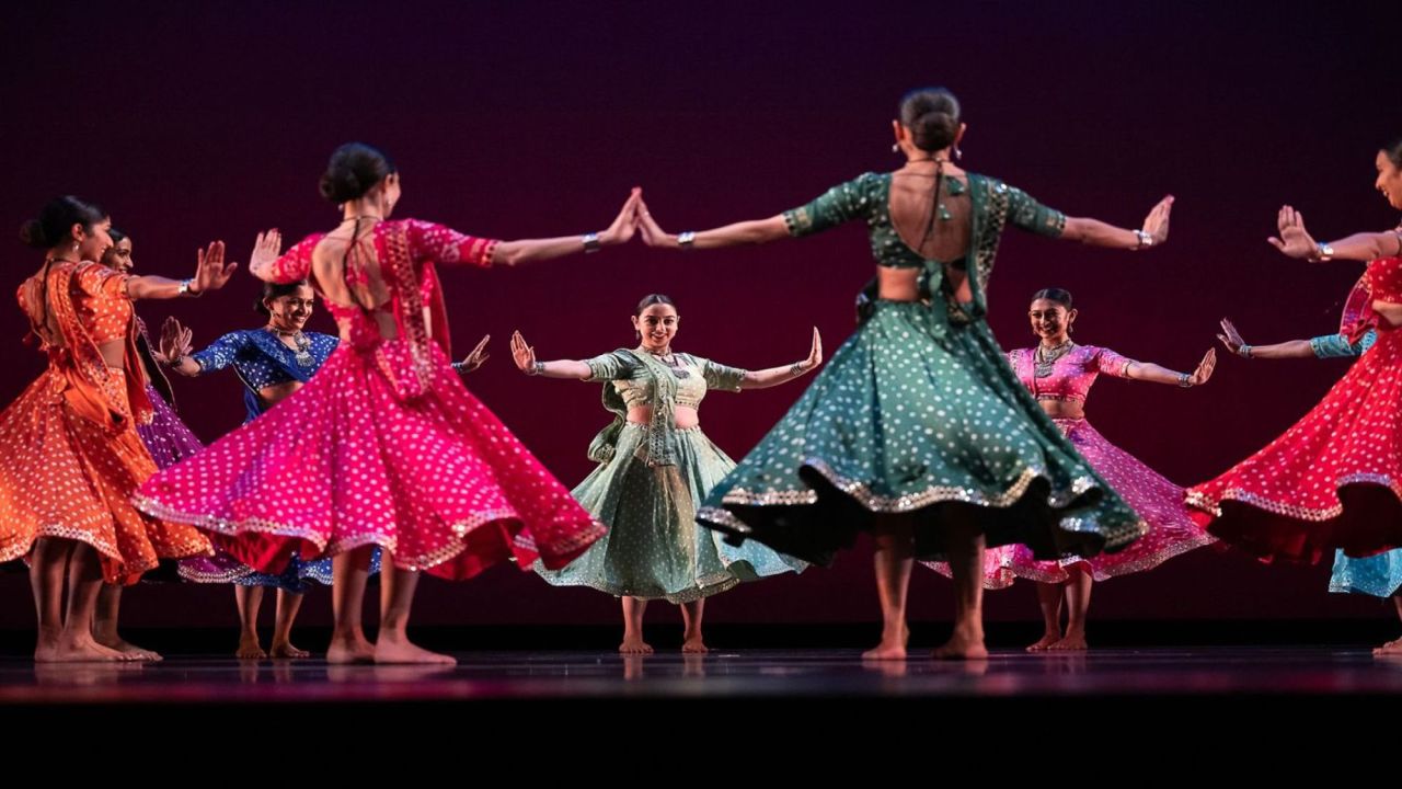 A group of dancers in colorful, traditional Indian attire perform in a circle on stage, their skirts swirling as they move with outstretched arms against a dark, purple-lit background. South Florida Business & Wealth