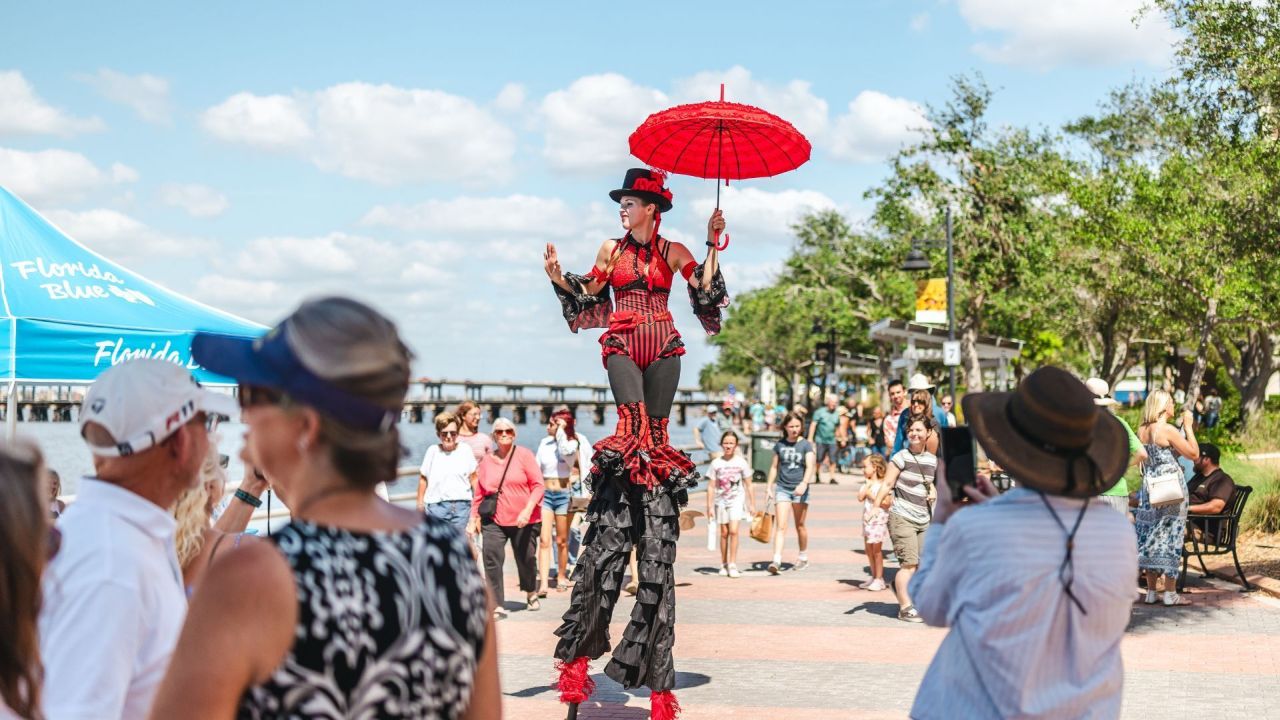 A performer on stilts wearing a red and black costume and holding a red umbrella entertains a crowd at an outdoor festival under a sunny sky. People walk and watch along the waterfront promenade. South Florida Business & Wealth
