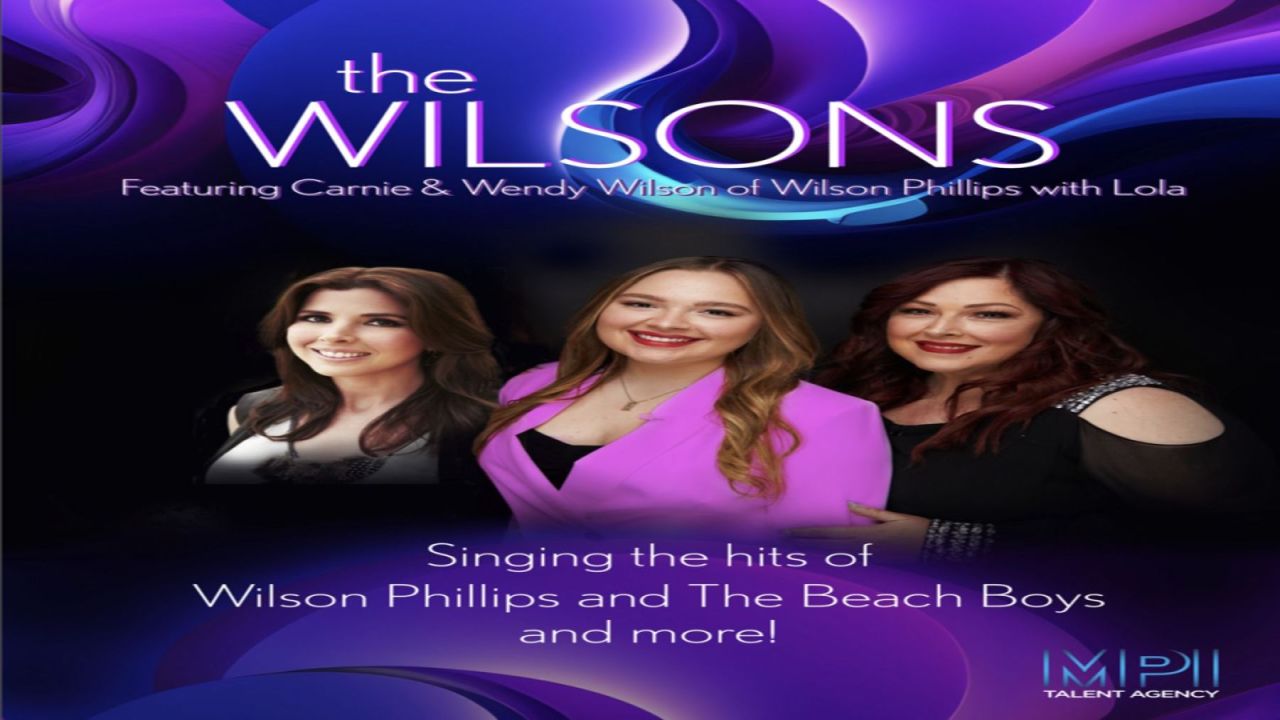 Three women—Carnie, Wendy, and Lola Wilson—stand smiling against a vibrant purple and blue abstract background. Text promotes their group, The Wilsons, performing hits from Wilson Phillips and The Beach Boys. South Florida Business & Wealth