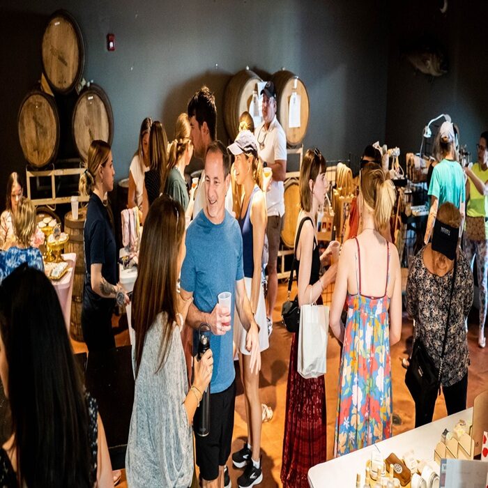 A lively group of people socialize and shop at an indoor market, surrounded by vendor tables and large wooden barrels against the wall. Some hold drinks and shopping bags while browsing products. South Florida Business & Wealth