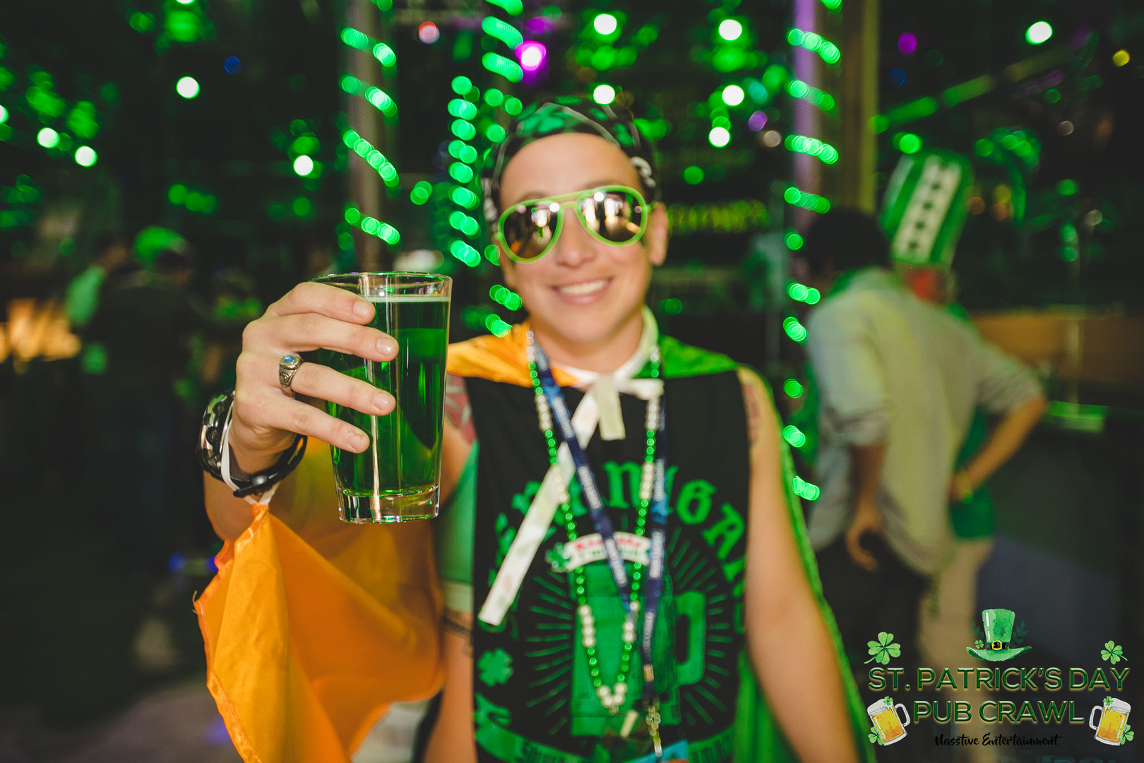 A person wearing sunglasses, a St. Patrick’s Day-themed outfit, and green beads holds up a glass of green drink at a festive, green-lit pub crawl event. Other people celebrate in the colorful background. South Florida Business & Wealth