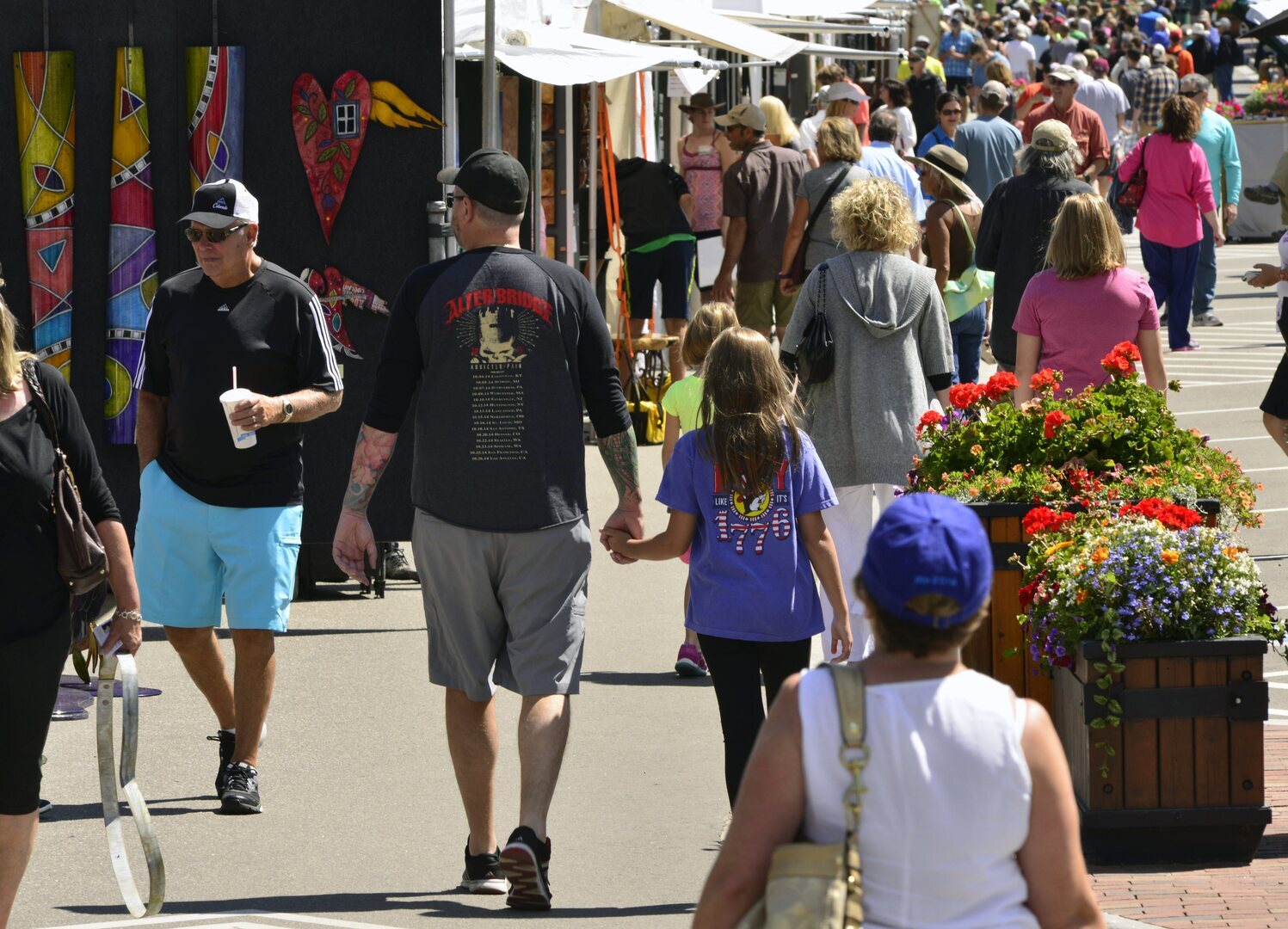 A busy outdoor art fair with people walking past colorful artwork and flower planters on a sunny day; families and individuals stroll along vendor booths displaying vibrant crafts. South Florida Business & Wealth