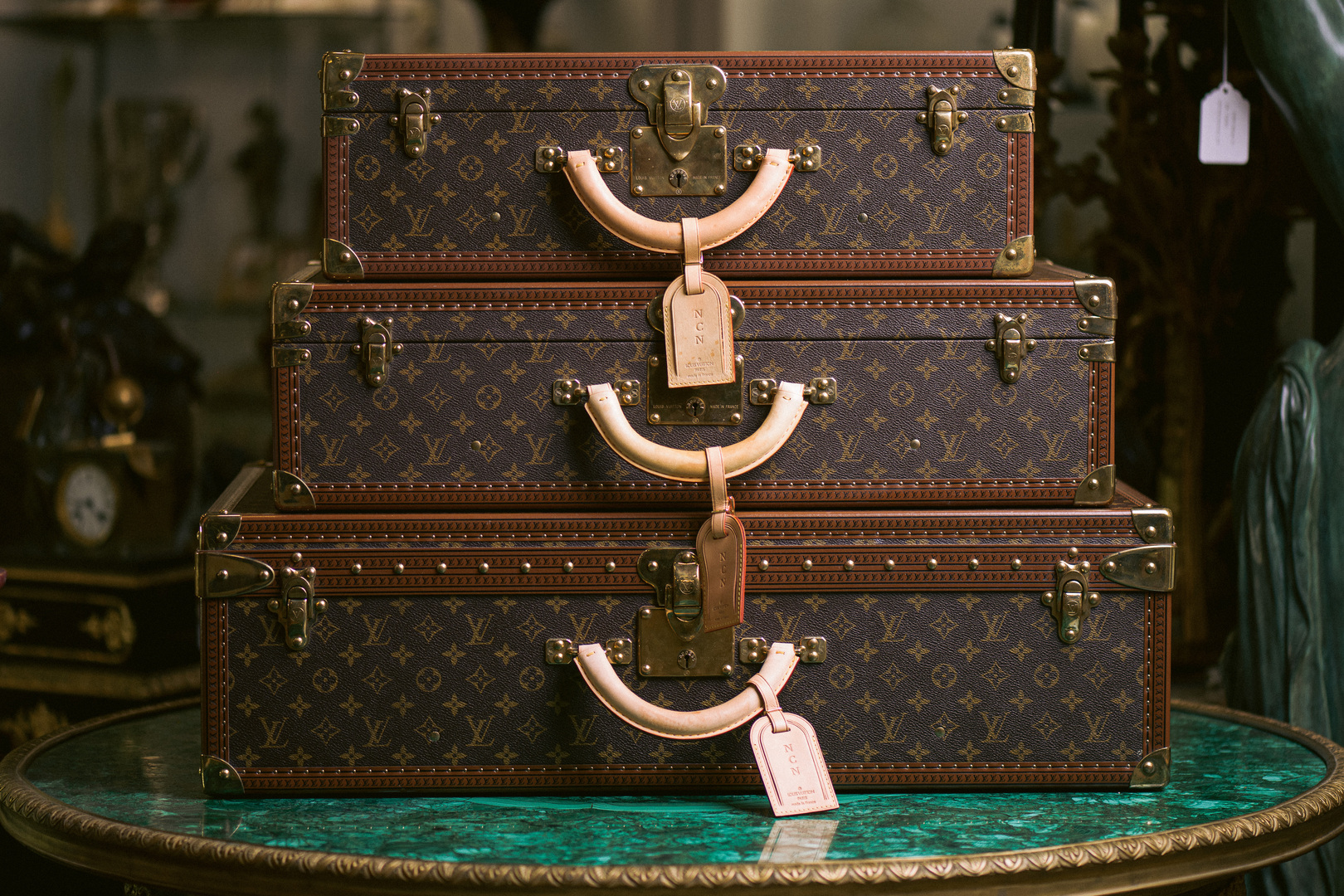 Three vintage Louis Vuitton suitcases with monogram pattern and leather handles are stacked on a round green marble table. Each suitcase has a tan luggage tag attached to the handle. South Florida Business & Wealth