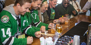 Five people wearing hockey jerseys and St. Patrick’s Day attire sit at a bar, smiling and preparing to play a game with wooden sticks and drinks in clear cups. South Florida Business & Wealth