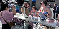 A group of people gathers at an outdoor table where a woman in sunglasses spins a prize wheel. Others, including a child, watch and participate. The table is covered with small items and containers under sunny weather. South Florida Business & Wealth