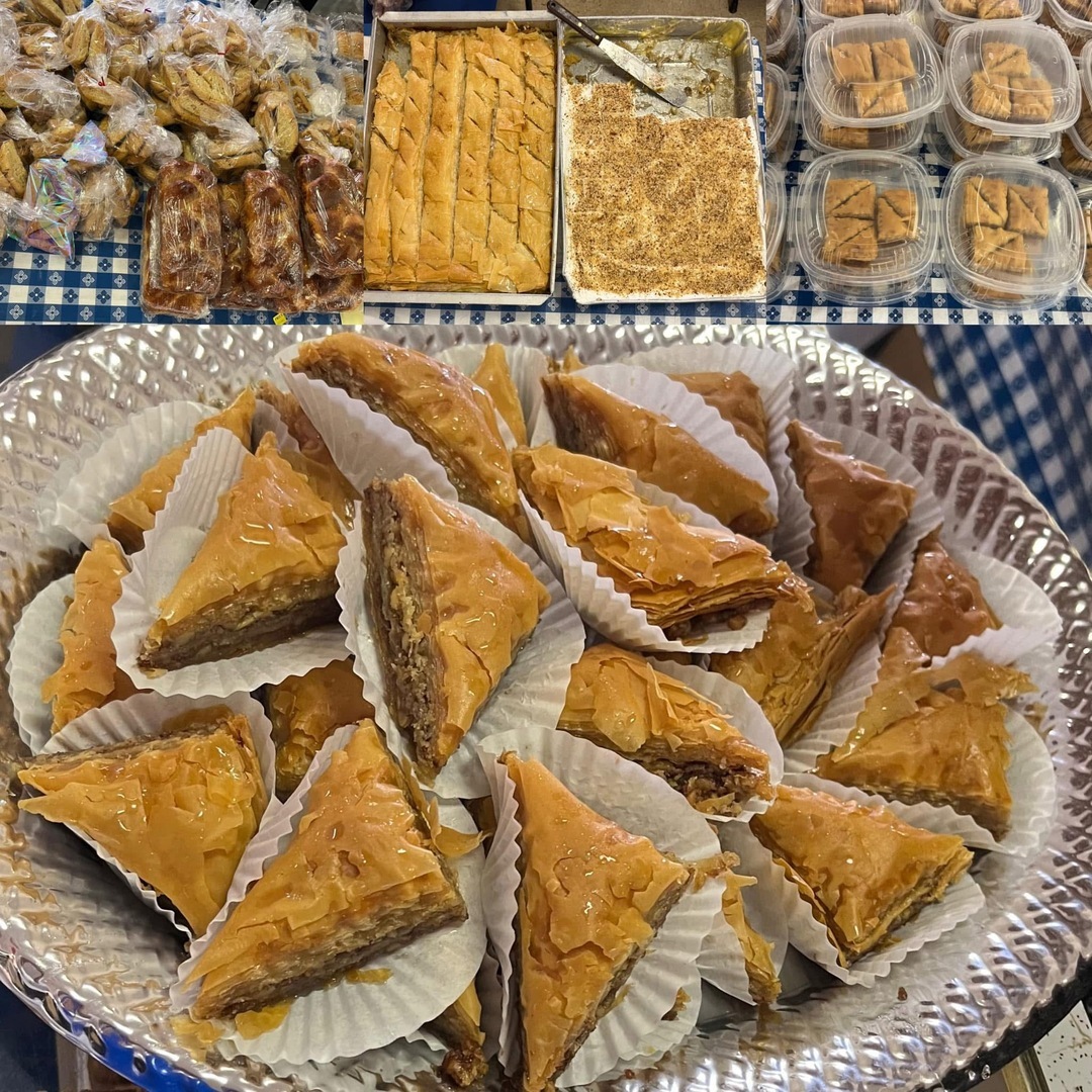 A platter of baklava triangles in white paper cups. Behind them are trays and containers filled with various types of Greek pastries on a checkered tablecloth. South Florida Business & Wealth