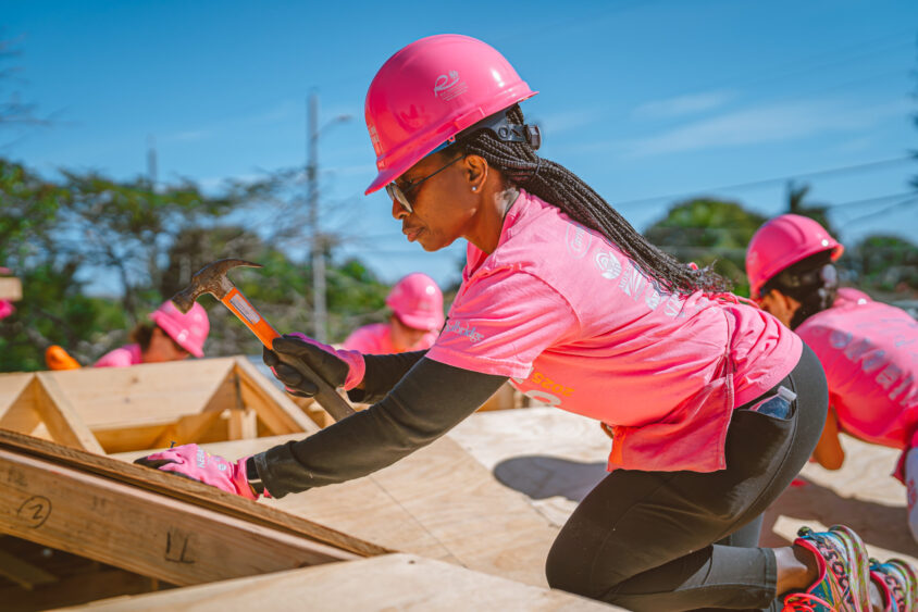 A person wearing a pink hard hat and shirt uses a hammer while working on a wooden structure outdoors, with others in similar attire working in the background. South Florida Business & Wealth