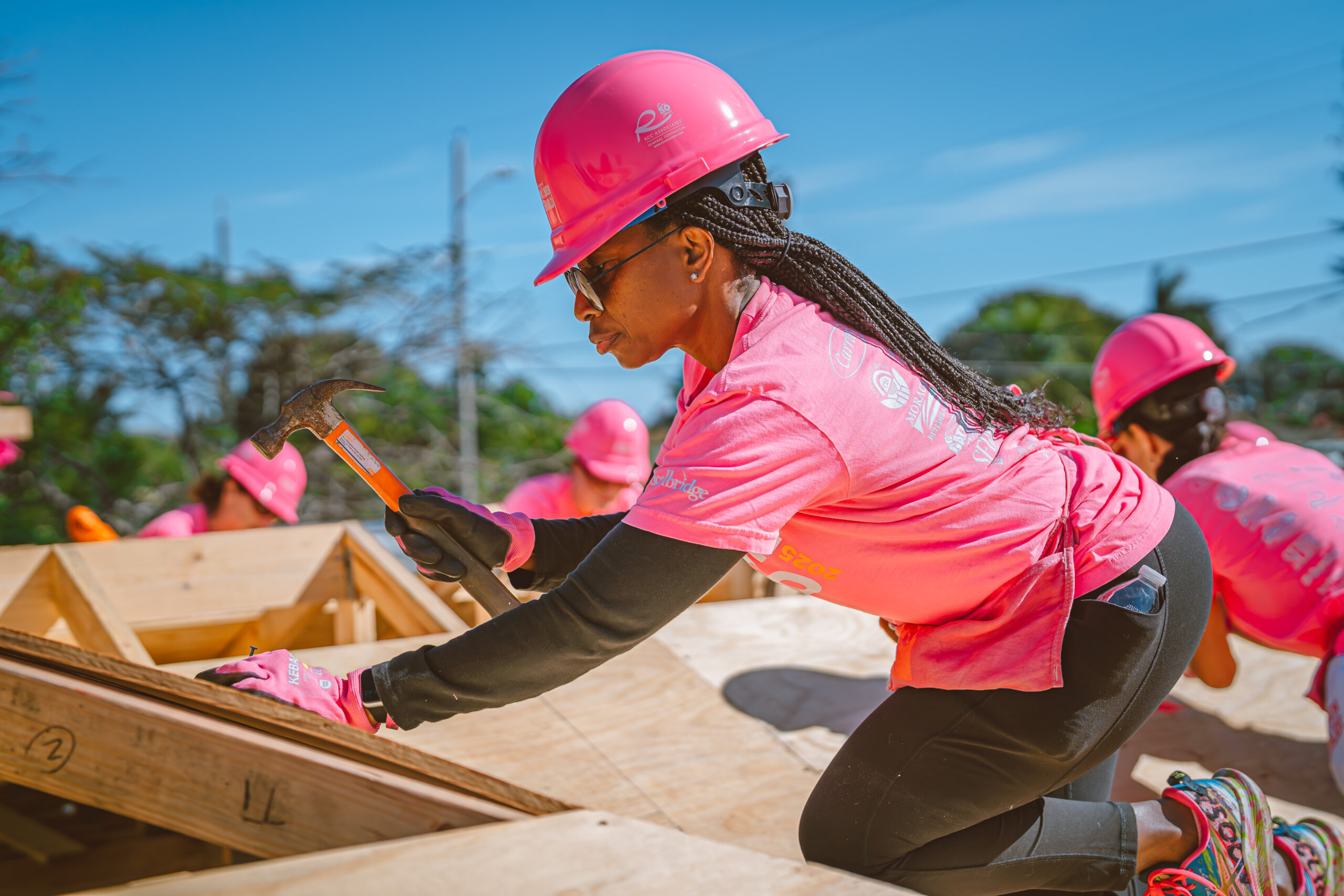 A person wearing a pink hard hat and shirt uses a hammer while working on a wooden structure outdoors, with others in similar attire working in the background. South Florida Business & Wealth