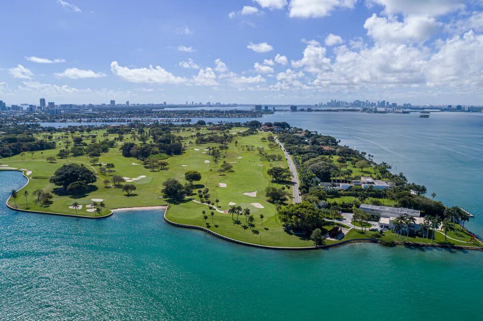 Aerial view of a green golf course on an island surrounded by blue water, with trees, sand traps, and several buildings, set against a city skyline in the background under a partly cloudy sky. South Florida Business & Wealth