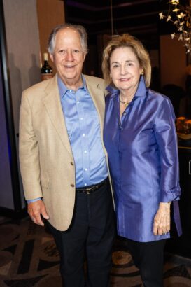 An older man in a tan blazer and blue shirt stands next to an older woman wearing a shiny blue tunic. They are smiling and posing together indoors at an event. South Florida Business & Wealth