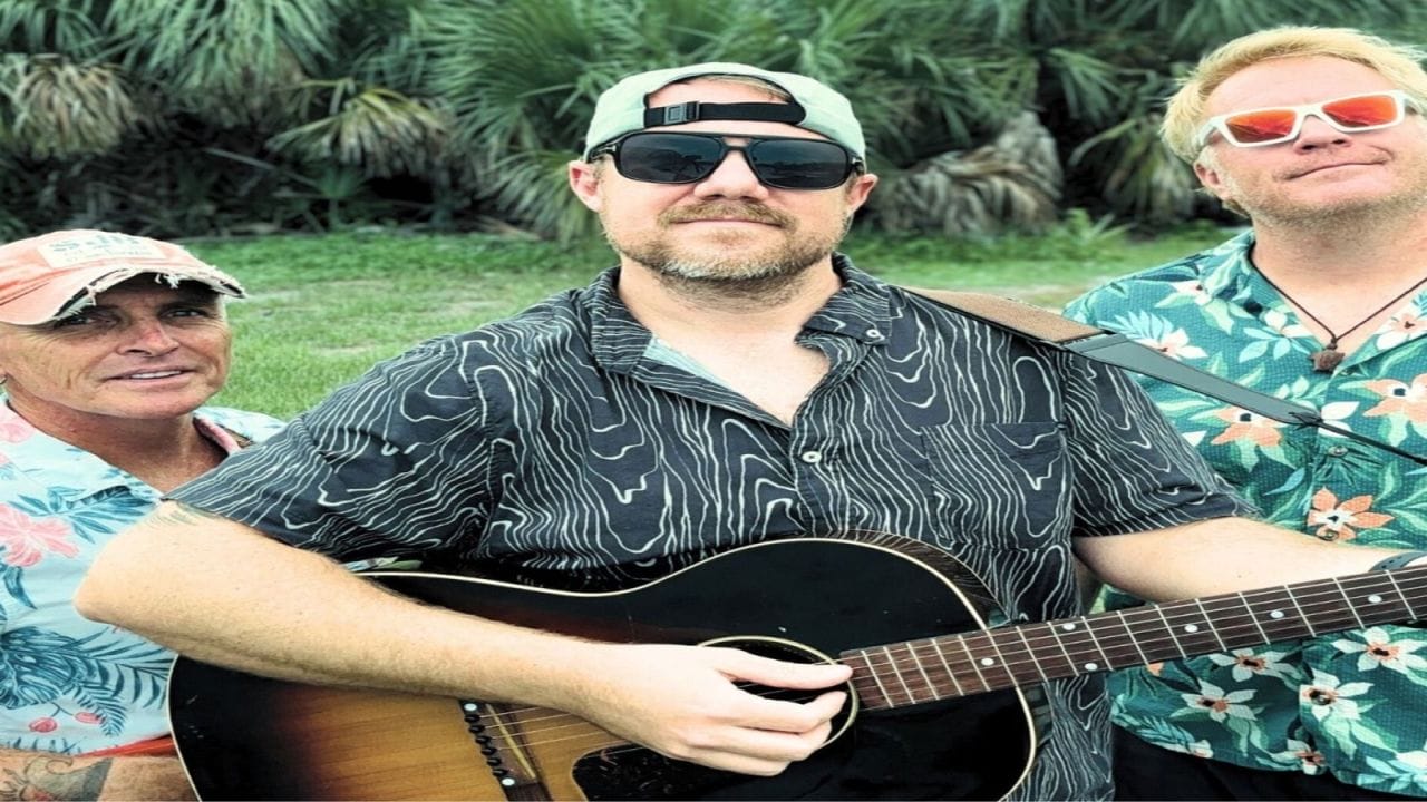 Three men in tropical shirts and sunglasses stand outdoors; the man in the center wears a backward cap and plays an acoustic guitar. Palm trees and greenery are visible in the background. South Florida Business & Wealth