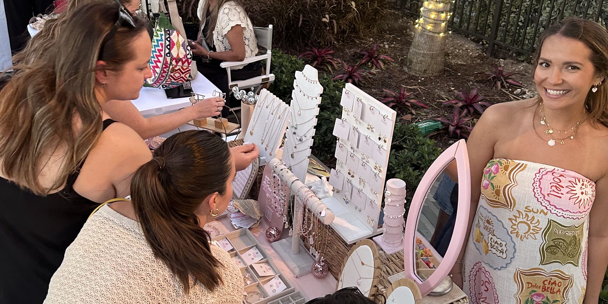 A group of women browse and try on jewelry at an outdoor market booth, which displays necklaces, earrings, and rings. One woman smiles at the camera; string lights and plants decorate the background. South Florida Business & Wealth