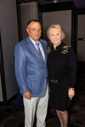 An older man in a blue suit and tie stands next to an older woman in a black dress with gold embellishments. They are both smiling and posing together indoors at a formal event. South Florida Business & Wealth